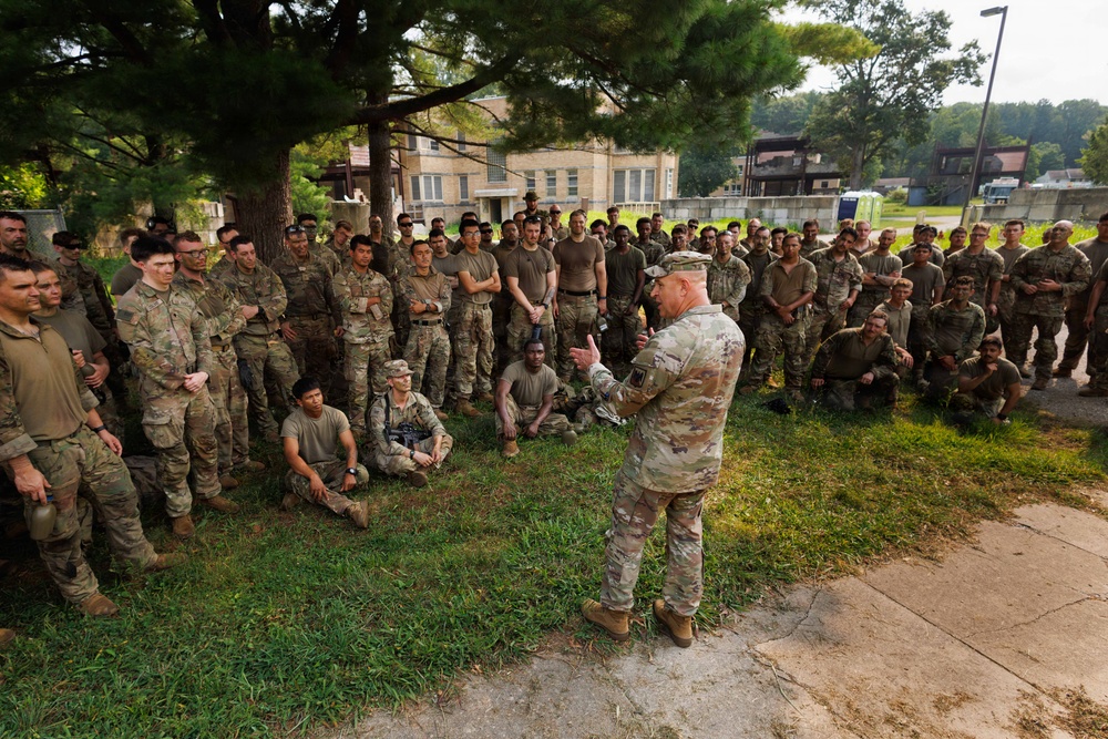A senior military officer in U.S. Army camouflage (OCP/MultiCam) uniform is addressing a large group of soldiers gathered outdoors near a tree on what appears to be a military installation. The soldiers are in various postures — standing, sitting, and kneeling — attentively listening to the briefing or address. The setting appears to be a training or garrison environment based on the surrounding buildings and infrastructure.