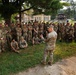 A senior military officer in U.S. Army camouflage (OCP/MultiCam) uniform is addressing a large group of soldiers gathered outdoors near a tree on what appears to be a military installation. The soldiers are in various postures — standing, sitting, and kneeling — attentively listening to the briefing or address. The setting appears to be a training or garrison environment based on the surrounding buildings and infrastructure.