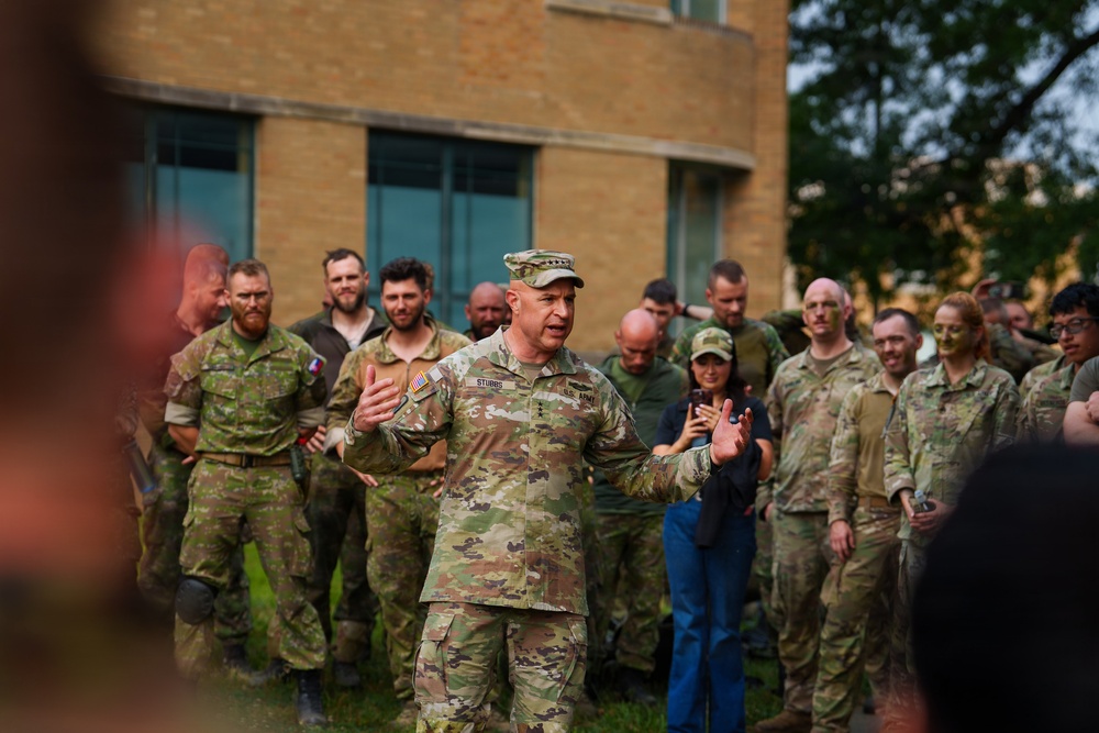 A U.S. Army officer with the name tape 'STUBBS' and four-star rank insignia is addressing a group of military personnel outdoors near a brick building. The assembled group includes soldiers in various camouflage uniforms, some with face paint applied, suggesting a recent or ongoing field exercise. The officer appears to be delivering a briefing or after-action remarks to a mixed multinational military audience.