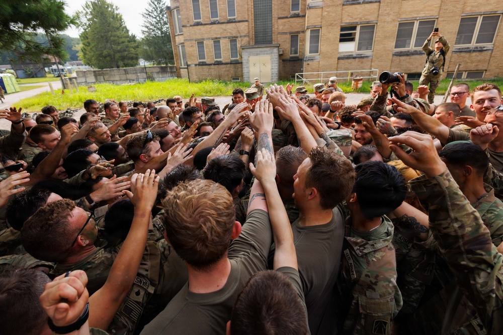 A large group of military personnel in camouflage uniforms are gathered in a team huddle, raising their hands together in a show of unit cohesion and morale. The scene takes place outdoors near a large brick building, likely a military installation or training facility. At least one individual on the upper right appears to be photographing or observing the event from an elevated position.