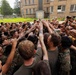 A large group of military personnel in camouflage uniforms are gathered in a team huddle, raising their hands together in a show of unit cohesion and morale. The scene takes place outdoors near a large brick building, likely a military installation or training facility. At least one individual on the upper right appears to be photographing or observing the event from an elevated position.