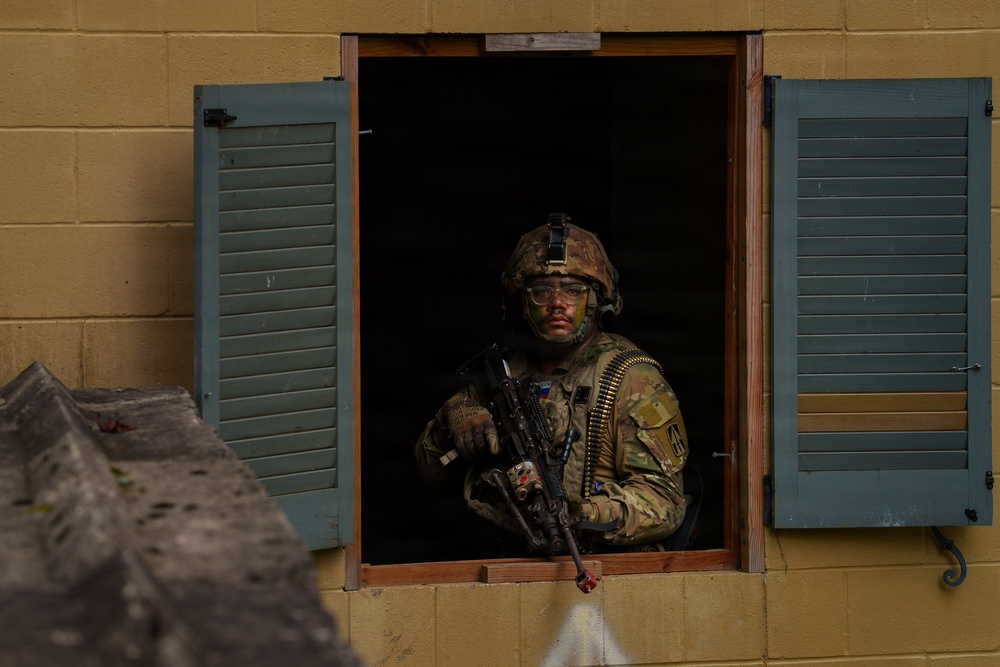 A fully equipped soldier in camouflage uniform and combat helmet is positioned at an open window of a cinder block building with teal/green shutters. The soldier is holding an automatic rifle and has a belt of ammunition draped across their body, with face paint applied. The setting appears to be a military training exercise or urban operations drill.