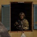 A fully equipped soldier in camouflage uniform and combat helmet is positioned at an open window of a cinder block building with teal/green shutters. The soldier is holding an automatic rifle and has a belt of ammunition draped across their body, with face paint applied. The setting appears to be a military training exercise or urban operations drill.
