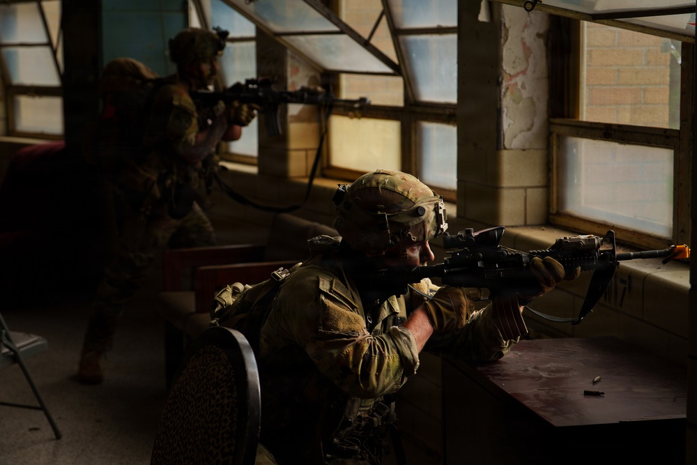 Two soldiers in full combat gear are positioned inside a building, aiming rifles through or toward windows. The foreground soldier is prone or crouched behind a desk, sighting down an assault rifle equipped with an optic. The setting appears to be a military training exercise conducted inside a structure resembling an office or classroom.