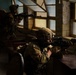 Two soldiers in full combat gear are positioned inside a building, aiming rifles through or toward windows. The foreground soldier is prone or crouched behind a desk, sighting down an assault rifle equipped with an optic. The setting appears to be a military training exercise conducted inside a structure resembling an office or classroom.