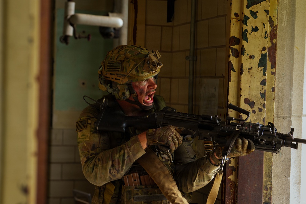 A soldier in full combat gear is positioned in a deteriorated building interior, holding a belt-fed machine gun and shouting. The soldier wears multicam camouflage, a tactical helmet with mounted accessories, and has face paint applied. The setting appears to be a military training exercise or operational environment inside a dilapidated structure.