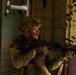 A soldier in full combat gear is positioned in a deteriorated building interior, holding a belt-fed machine gun and shouting. The soldier wears multicam camouflage, a tactical helmet with mounted accessories, and has face paint applied. The setting appears to be a military training exercise or operational environment inside a dilapidated structure.