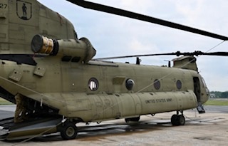 The image shows a United States Army CH-47 Chinook tandem-rotor heavy-lift helicopter parked on a tarmac or airfield. The aircraft is painted in standard olive drab/army green livery with 'UNITED STATES ARMY' markings visible on the fuselage. The rear cargo ramp appears to be lowered, and at least one soldier in camouflage uniform is visible near the aircraft.