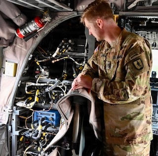 A U.S. Army soldier in OCP (Operational Camouflage Pattern) uniform is performing maintenance on a military aircraft, examining an open access panel revealing complex wiring, hydraulic lines, and mechanical components. The soldier appears to be inspecting or servicing the interior systems of what appears to be a rotary-wing aircraft. A red fire extinguisher is visible mounted near the access panel.
