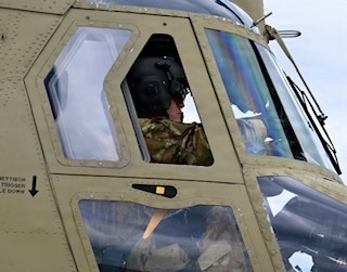A military helicopter pilot or crew member is seated inside a olive-drab/tan colored military helicopter cockpit. The individual is wearing a flight helmet with communications equipment and military camouflage uniform. The image is taken from an exterior angle looking up at the cockpit windows.