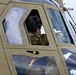 A military helicopter pilot or crew member is seated inside a olive-drab/tan colored military helicopter cockpit. The individual is wearing a flight helmet with communications equipment and military camouflage uniform. The image is taken from an exterior angle looking up at the cockpit windows.