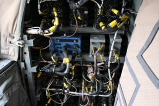 The image shows an open access panel or bay of what appears to be a military or large commercial aircraft, revealing a dense array of hydraulic lines, electrical wiring, and avionics components. Numerous yellow-capped connectors and fittings are prominently visible throughout the assembly, along with a blue electronic module mounted centrally. The area appears to be an avionics or systems bay, likely photographed during maintenance or inspection.