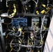 The image shows an open access panel or bay of what appears to be a military or large commercial aircraft, revealing a dense array of hydraulic lines, electrical wiring, and avionics components. Numerous yellow-capped connectors and fittings are prominently visible throughout the assembly, along with a blue electronic module mounted centrally. The area appears to be an avionics or systems bay, likely photographed during maintenance or inspection.