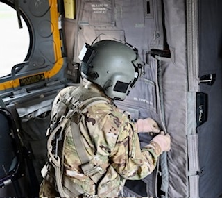 A military personnel member is seated inside what appears to be a military helicopter, viewed from behind. The individual is wearing full combat gear including a flight helmet with mounted equipment, multicam/OCP pattern uniform, and tactical vest with harness straps. They appear to be interacting with interior door or wall components of the aircraft.