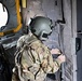 A military personnel member is seated inside what appears to be a military helicopter, viewed from behind. The individual is wearing full combat gear including a flight helmet with mounted equipment, multicam/OCP pattern uniform, and tactical vest with harness straps. They appear to be interacting with interior door or wall components of the aircraft.