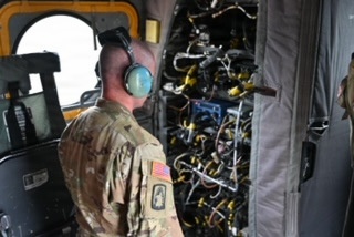A U.S. Army soldier wearing camouflage uniform and aviation headset is inspecting or examining a complex wiring and avionics panel inside what appears to be a military helicopter. The soldier is viewed from behind, facing an exposed panel filled with dense wiring harnesses and yellow-tagged components. The setting appears to be the interior of a large military rotary-wing aircraft undergoing maintenance or inspection.