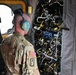 A U.S. Army soldier wearing camouflage uniform and aviation headset is inspecting or examining a complex wiring and avionics panel inside what appears to be a military helicopter. The soldier is viewed from behind, facing an exposed panel filled with dense wiring harnesses and yellow-tagged components. The setting appears to be the interior of a large military rotary-wing aircraft undergoing maintenance or inspection.