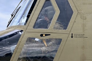 The image shows the cockpit area of a military helicopter, painted in olive drab/tan color, with at least one occupant visible wearing a flight helmet and military uniform. The exterior of the aircraft shows riveted paneling and a door jettison instruction label. The photograph appears to have been taken from outside the aircraft at close range.