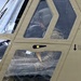 The image shows the cockpit area of a military helicopter, painted in olive drab/tan color, with at least one occupant visible wearing a flight helmet and military uniform. The exterior of the aircraft shows riveted paneling and a door jettison instruction label. The photograph appears to have been taken from outside the aircraft at close range.