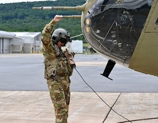 A military personnel in full combat uniform and flight helmet is conducting a pre-flight or ground inspection of a military helicopter on a tarmac. The individual has one arm raised, possibly signaling or inspecting the rotor blade area, while holding equipment in the other hand. The setting appears to be a military airfield with hangar structures and tree-covered hills visible in the background.