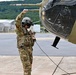 A military personnel in full combat uniform and flight helmet is conducting a pre-flight or ground inspection of a military helicopter on a tarmac. The individual has one arm raised, possibly signaling or inspecting the rotor blade area, while holding equipment in the other hand. The setting appears to be a military airfield with hangar structures and tree-covered hills visible in the background.