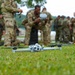 A military drone (quadcopter) sits on a grass field in the foreground while a group of soldiers in camouflage uniforms gather in the background. One soldier appears to be operating or inspecting a controller device. The scene appears to be a training exercise or demonstration involving unmanned aerial vehicle (UAV) operations.