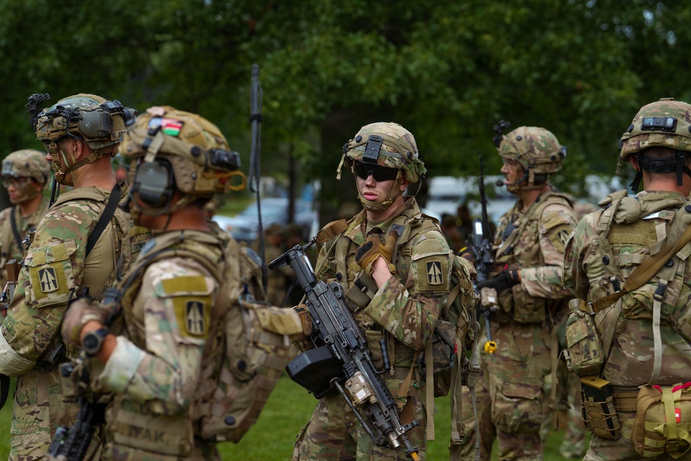 A group of heavily equipped military soldiers in MultiCam/OCP camouflage uniforms are assembled in an outdoor area with green trees in the background. The soldiers are wearing full combat gear including helmets with night vision mounts, body armor, and tactical accessories. At least one soldier is visibly holding an assault rifle, and unit patches are visible on their shoulders.