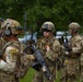 A group of heavily equipped military soldiers in MultiCam/OCP camouflage uniforms are assembled in an outdoor area with green trees in the background. The soldiers are wearing full combat gear including helmets with night vision mounts, body armor, and tactical accessories. At least one soldier is visibly holding an assault rifle, and unit patches are visible on their shoulders.