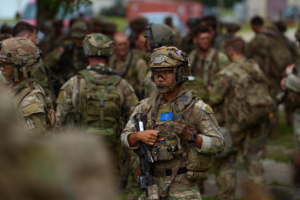 A group of military personnel in full combat gear are assembled in an outdoor area, likely during a training exercise or operational briefing. The central figure is a soldier wearing MultiCam camouflage, a tactical helmet with goggles, a communications headset, and carrying a rifle. A blue patch with a yellow star pattern — consistent with an Indiana National Guard state flag patch — is visible on the central soldier's chest.