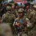 A group of military personnel in full combat gear are assembled in an outdoor area, likely during a training exercise or operational briefing. The central figure is a soldier wearing MultiCam camouflage, a tactical helmet with goggles, a communications headset, and carrying a rifle. A blue patch with a yellow star pattern — consistent with an Indiana National Guard state flag patch — is visible on the central soldier's chest.