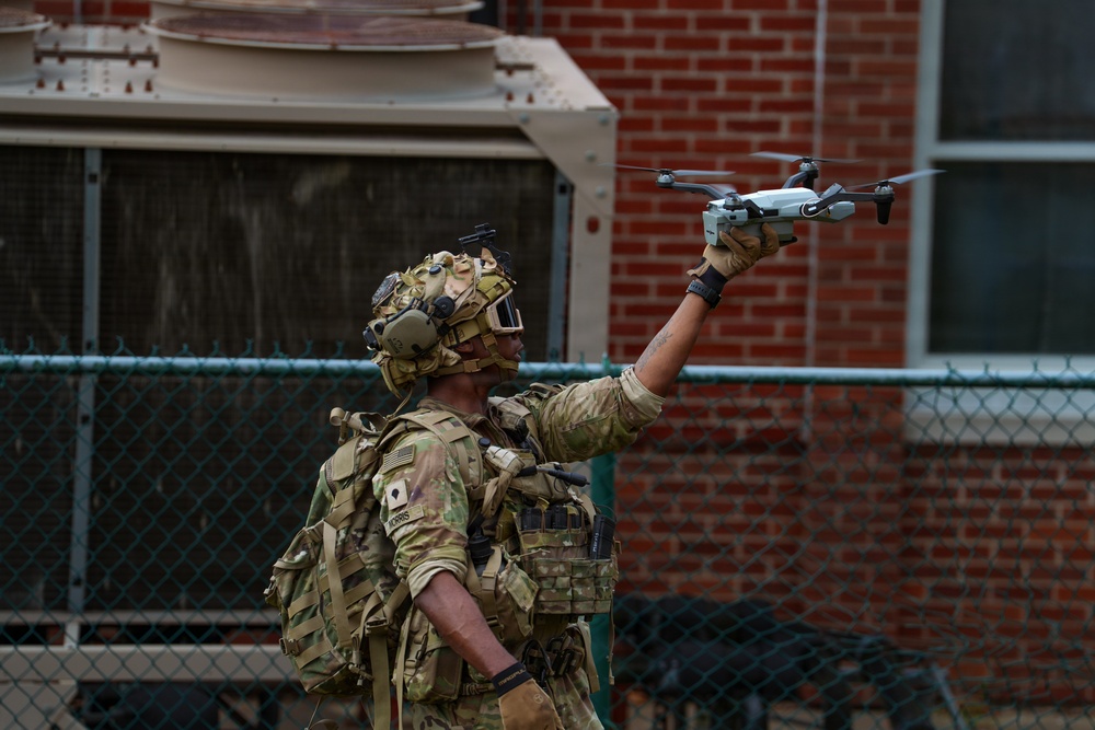 A heavily equipped military or special operations soldier is holding a small quadcopter drone (appears to be a DJI Spark or similar consumer-grade drone) aloft with one hand, likely preparing to launch it. The soldier is wearing full tactical gear including a multicam uniform, plate carrier, tactical helmet with night vision mounts and communications headset, and a backpack. The setting appears to be an urban training environment near a brick building with a chain-link fence.