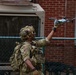 A heavily equipped military or special operations soldier is holding a small quadcopter drone (appears to be a DJI Spark or similar consumer-grade drone) aloft with one hand, likely preparing to launch it. The soldier is wearing full tactical gear including a multicam uniform, plate carrier, tactical helmet with night vision mounts and communications headset, and a backpack. The setting appears to be an urban training environment near a brick building with a chain-link fence.