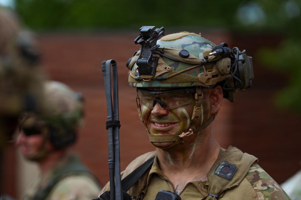 A military soldier in full tactical gear is shown in close-up, wearing camouflage face paint in green and black tones. The soldier is equipped with an advanced combat helmet featuring multiple attachments including night vision mount brackets, communications headset, and a camera or sensor device. A second soldier is visible in the blurred background near a brick wall.