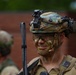 A military soldier in full tactical gear is shown in close-up, wearing camouflage face paint in green and black tones. The soldier is equipped with an advanced combat helmet featuring multiple attachments including night vision mount brackets, communications headset, and a camera or sensor device. A second soldier is visible in the blurred background near a brick wall.