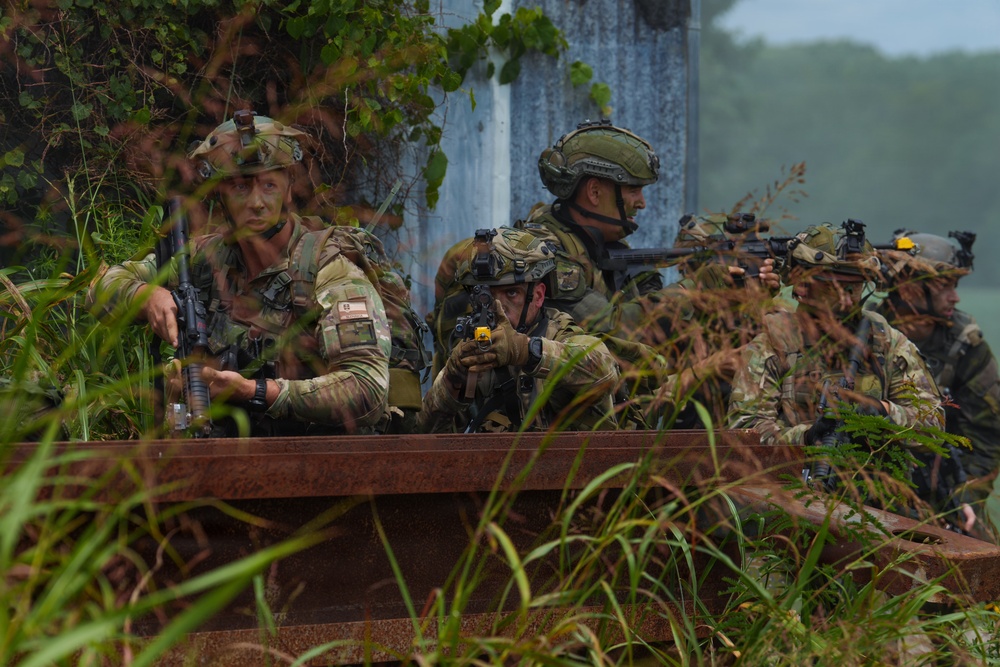 A group of approximately five soldiers in camouflage uniforms are taking cover behind a rusted metal barrier or rail in a vegetated, outdoor environment. They are in tactical positions, with weapons raised and aimed, suggesting a military training exercise or field operation. The soldiers wear various helmet configurations and face camouflage paint, indicating a coordinated unit.