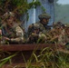 A group of approximately five soldiers in camouflage uniforms are taking cover behind a rusted metal barrier or rail in a vegetated, outdoor environment. They are in tactical positions, with weapons raised and aimed, suggesting a military training exercise or field operation. The soldiers wear various helmet configurations and face camouflage paint, indicating a coordinated unit.