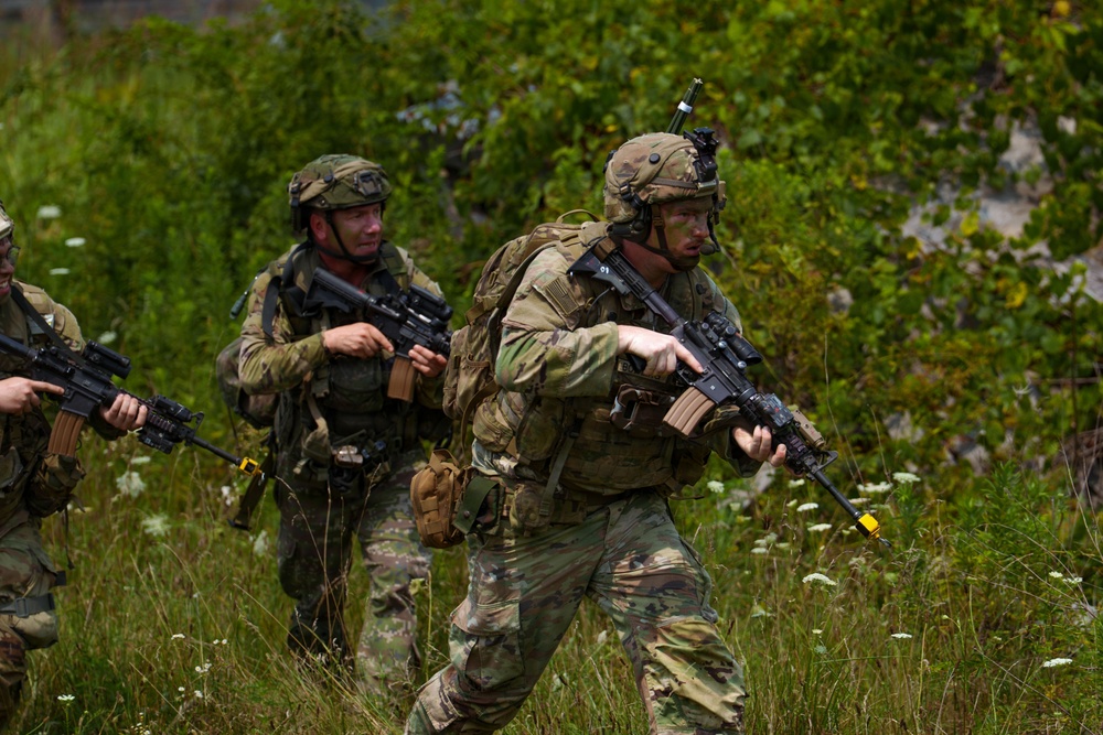 The image shows a group of military personnel in full combat gear moving through a vegetated outdoor terrain. They are equipped with assault rifles, tactical helmets, body armor, and camouflage uniforms. The presence of yellow/orange barrel markers on the weapons suggests this is a training exercise rather than live combat.