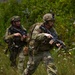 The image shows a group of military personnel in full combat gear moving through a vegetated outdoor terrain. They are equipped with assault rifles, tactical helmets, body armor, and camouflage uniforms. The presence of yellow/orange barrel markers on the weapons suggests this is a training exercise rather than live combat.