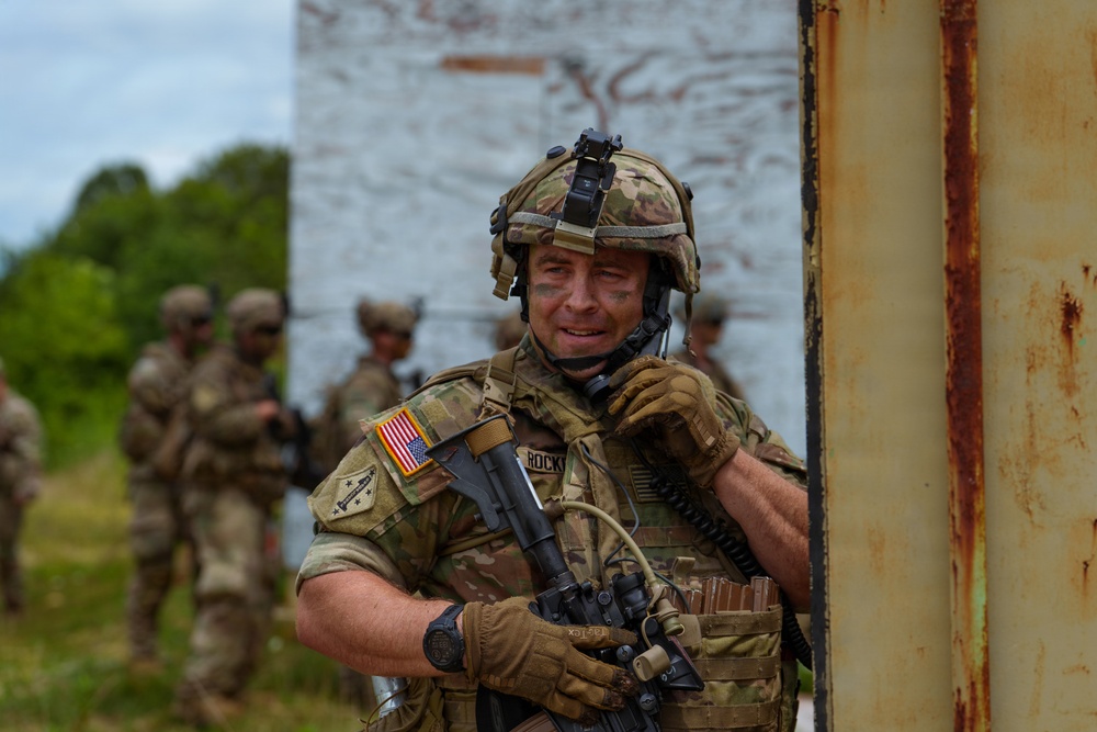 A U.S. Army soldier in full combat gear is positioned near a rusted metal structure, appearing to be engaged in a field training exercise. The soldier wears OCP (Operational Camouflage Pattern) uniform with an American flag patch and a name tape reading 'ROCKY', and has face camouflage paint applied. Additional soldiers in similar gear are visible in the blurred background.