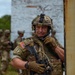 A U.S. Army soldier in full combat gear is positioned near a rusted metal structure, appearing to be engaged in a field training exercise. The soldier wears OCP (Operational Camouflage Pattern) uniform with an American flag patch and a name tape reading 'ROCKY', and has face camouflage paint applied. Additional soldiers in similar gear are visible in the blurred background.