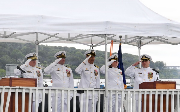 A formal U.S. Navy military ceremony is taking place on an outdoor platform or pier decorated with red, white, and blue bunting. Five naval officers in white dress uniforms are standing at attention, with four of them rendering salutes. The setting appears to be waterside, with a body of water, trees, and a bridge visible in the background.