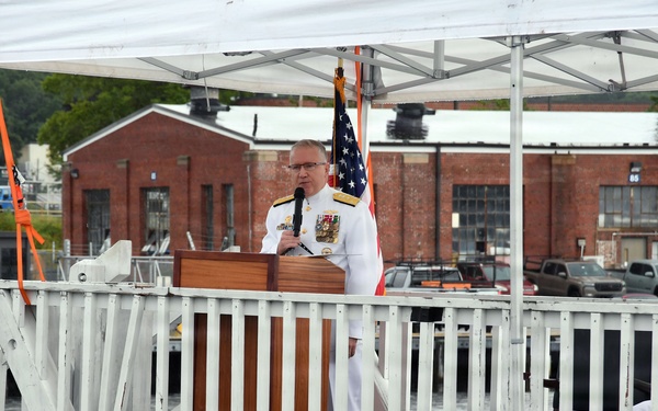 A naval officer in white dress uniform delivers a speech at a podium during what appears to be an official military ceremony at a waterfront facility. The event is held on a dock or pier platform decorated with red, white, and blue bunting, with an American flag displayed behind the speaker. A second uniformed officer is seated to the right, and brick buildings consistent with a naval installation are visible in the background.