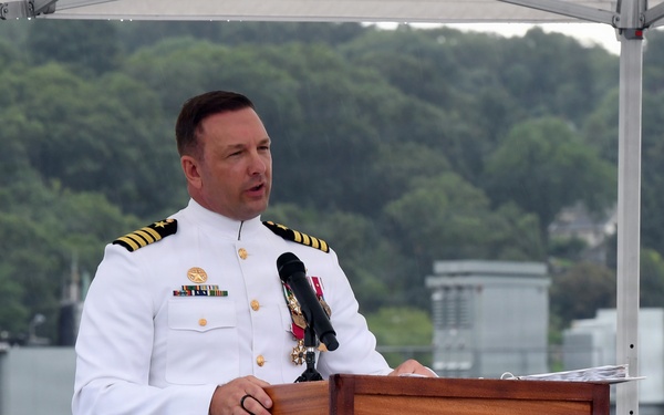 A naval officer in white dress uniform is speaking at a wooden podium during what appears to be an outdoor military ceremony. The setting appears to be on or near a waterfront, with a white railing visible and a canopy overhead. The officer displays multiple medals and rank insignia consistent with a senior officer.