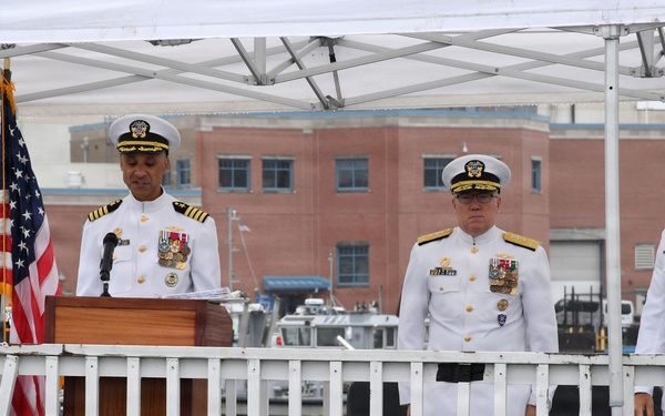Three U.S. Navy officers in white dress uniforms are standing on an elevated platform or vessel deck during what appears to be a formal military ceremony. One officer is speaking at a wooden podium with a microphone, while two others stand at attention behind a white railing. An American flag is displayed to the left of the podium.