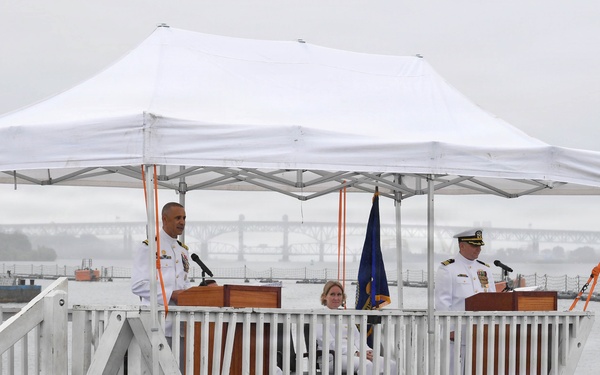 A formal U.S. Navy ceremony is taking place on an elevated outdoor platform near a waterfront, likely a change of command or commissioning ceremony. Two naval officers in white dress uniforms stand at separate podiums under a white canopy tent, with a civilian woman seated between them. The platform is decorated with red, white, and blue bunting, and both an American flag and a Navy flag are visible.