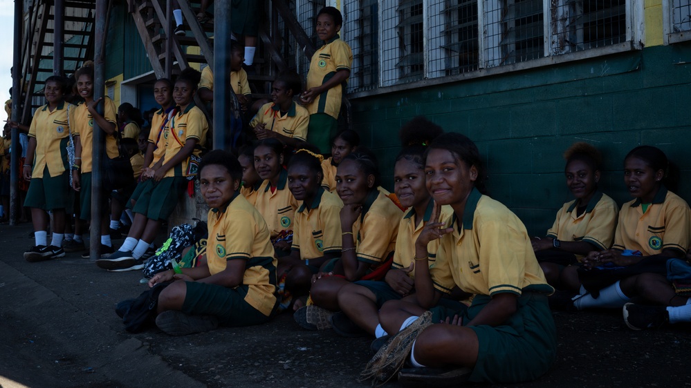 A large group of students wearing matching yellow and green school uniforms are gathered along a wall, with some sitting on the ground and others standing or elevated on a structure. The setting appears to be an outdoor area adjacent to a green-painted building with barred windows. The students appear to be waiting or observing an event.