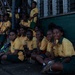 A large group of students wearing matching yellow and green school uniforms are gathered along a wall, with some sitting on the ground and others standing or elevated on a structure. The setting appears to be an outdoor area adjacent to a green-painted building with barred windows. The students appear to be waiting or observing an event.