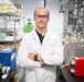 A male laboratory professional wearing a white lab coat and safety glasses stands with arms crossed in what appears to be a well-equipped research or industrial laboratory. He is posed confidently in front of complex equipment including tubing, pumps, and instrumentation. The setting suggests a chemical, biotech, or engineering research environment.