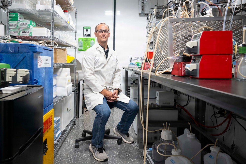 A male researcher or scientist is seated on a lab stool in a well-equipped laboratory setting, wearing a white lab coat, safety glasses, and jeans. He is posed calmly with hands clasped, surrounded by laboratory equipment, shelving units, tubing, and chemical storage containers. The image appears to be a professional portrait taken in an active research laboratory.