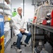 A male researcher or scientist is seated on a lab stool in a well-equipped laboratory setting, wearing a white lab coat, safety glasses, and jeans. He is posed calmly with hands clasped, surrounded by laboratory equipment, shelving units, tubing, and chemical storage containers. The image appears to be a professional portrait taken in an active research laboratory.